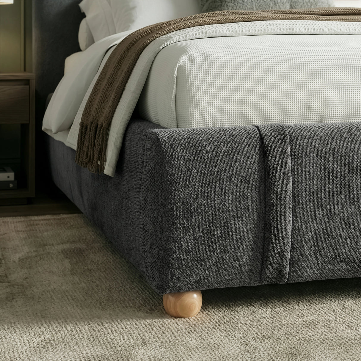 Lower base corner of king-size bed frame in dark grey upholstery with a rounded edge and short wooden legs