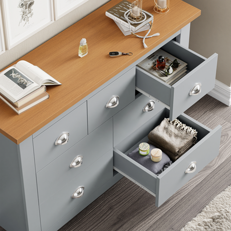 7 Drawer Wide Chest of Drawers with a wooden top and grey body, featuring silver cup handles and open drawers showing folded clothes and accessories.
