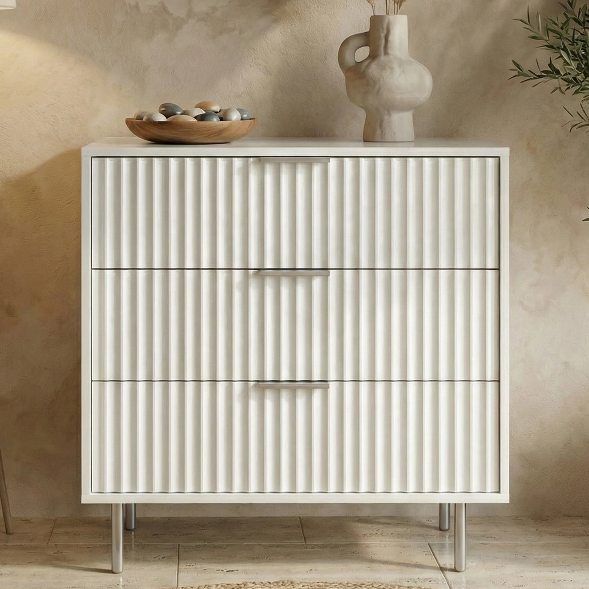 White Flutrina 3 drawer chest with vertical panels and slim metal legs in a bedroom setting