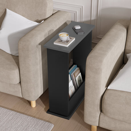 Black side table with a cup and books next to beige armchairs in a living room.