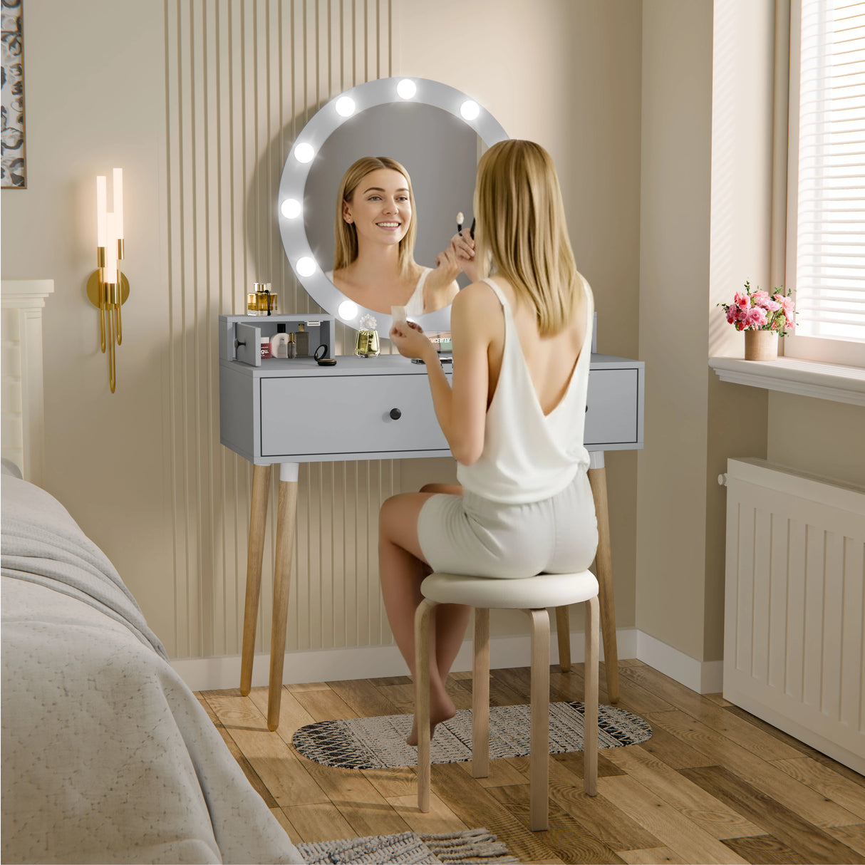 Woman applying makeup in front of a vanity mirror with lights in a bedroom setting.