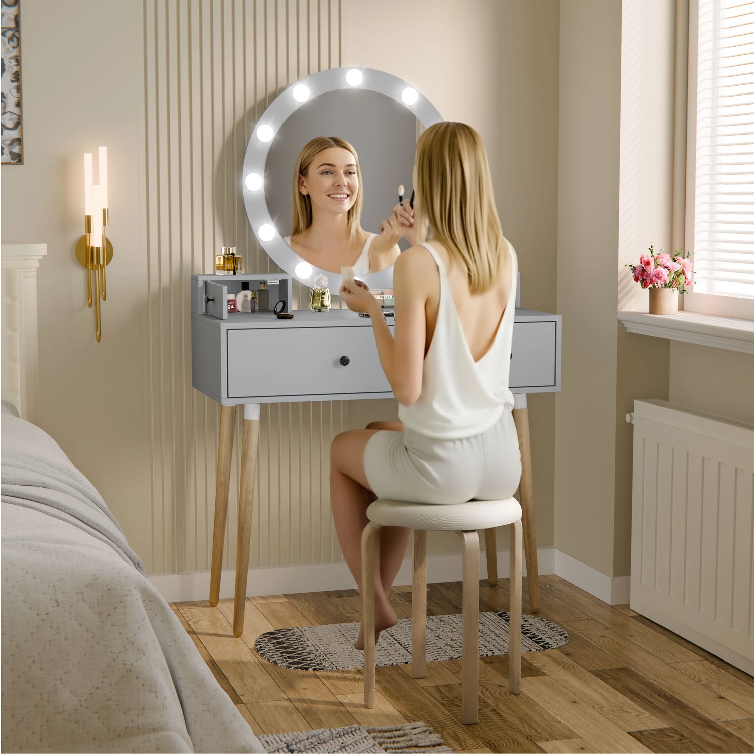 Woman applying makeup in front of a vanity mirror with lights in a bedroom setting.
