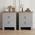 [Grey] Pair of grey bedside table pair on rug, oak-effect tops, chrome cup handles, lamp and books left, vase and frame right