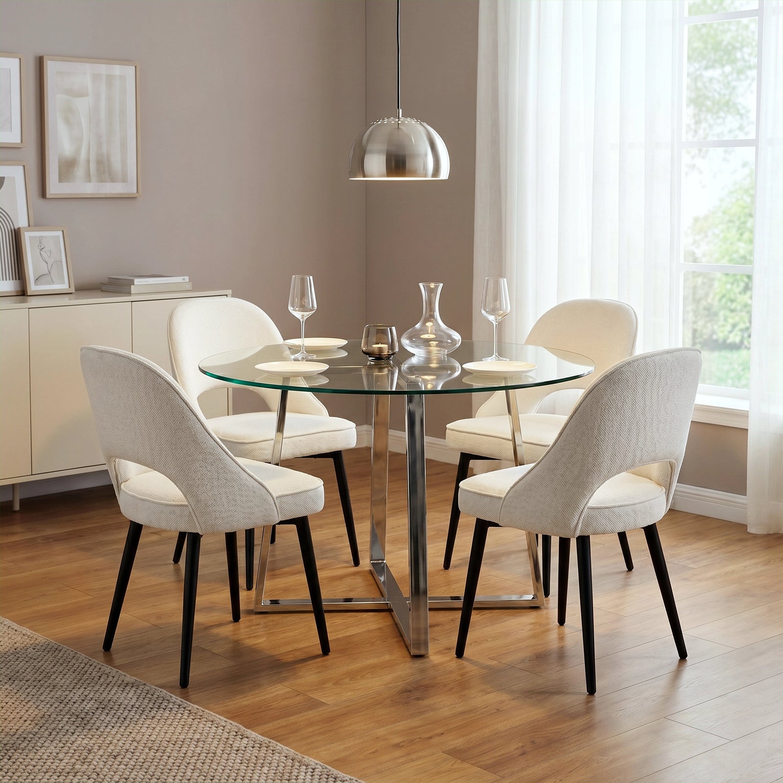 [Clear] 4-seater round dining table with a clear glass top, chrome base, styled with cream chairs, plates, glasses, & candle.
