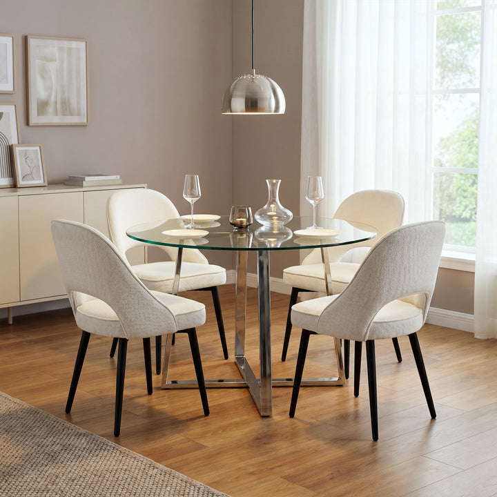 [Clear] 4-seater round dining table with a clear glass top, chrome base, styled with cream chairs, plates, glasses, & candle.

