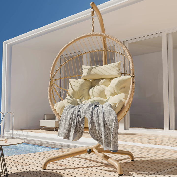 [Beige] Side view of beige hanging egg chair on wood deck, pool to the left, cream cushions stacked