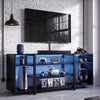 [Black Carbon Fibre] Zelumi TV stand front-facing in a brick-wall gaming room with blue LED shelves, console, VR headset, soundbar, and items on top