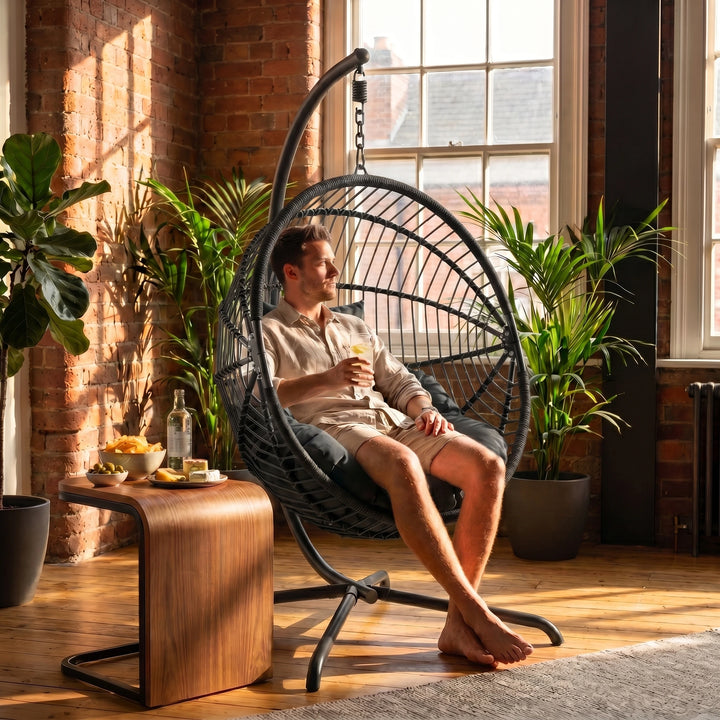 [Black] Man relaxing in a hanging egg chair in a sunlit room with plants and a wooden table