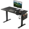 [110cm x 60cm] Electric Standing Desk Height Adjustable featuring an all-black design with a computer monitor, integrated desk mat, hanging headphones, and a grey fabric side storage pocket