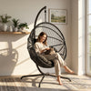 [Black] Woman sitting in a black hanging chair with a cat, in a bright room with plants and a painting