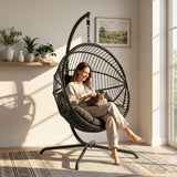 [Black] Woman sitting in a black hanging chair with a cat, in a bright room with plants and a painting