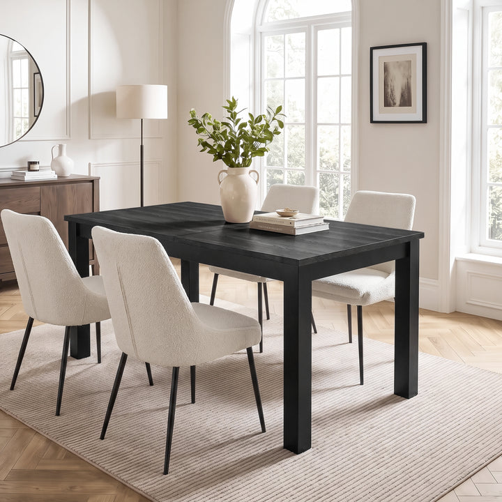 [140cm | Black] Closed dining table with four cream chairs, black wood top, leafy vase, books, dish and black frame.