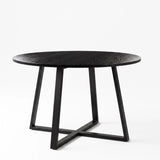 [Black Wood] Round wood-effect dining table in a studio view with the tabletop resting above a black iron cross base.