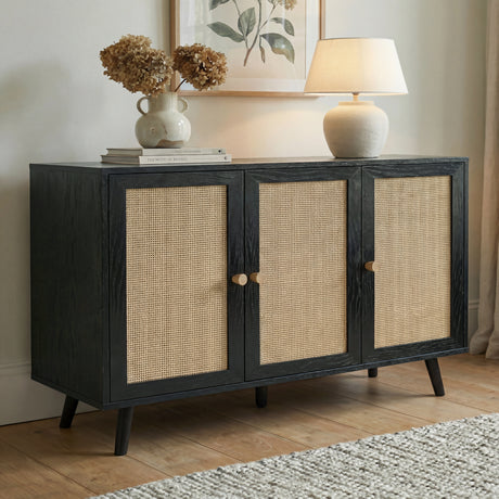 [Black] Wovena 3-door sideboard in an angled lifestyle view on oak flooring below a botanical print, topped with dried hydrangeas in a white vase and a round table lamp