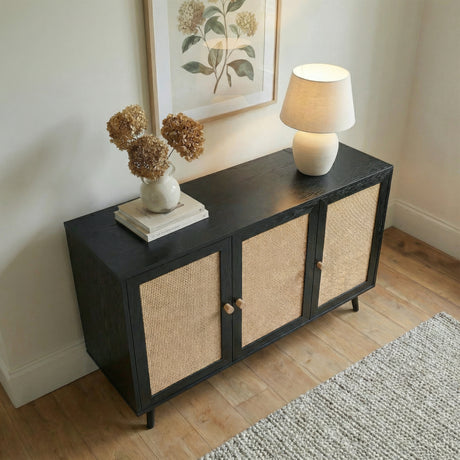 [Black] Wovena 3-door sideboard seen from above at an angle in a hallway setting, styled with a round white vase of dried hydrangeas and a ceramic table lamp to the right