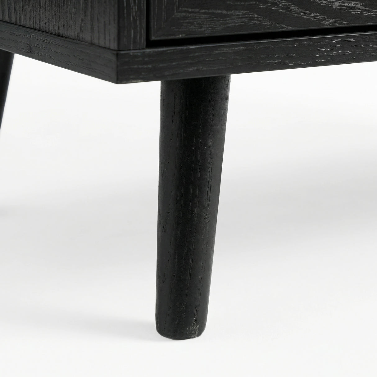 [Black] Wovena close-up of a solid tapered black-stained leg showing natural wood grain through the dark finish where it meets the ebonised cabinet base