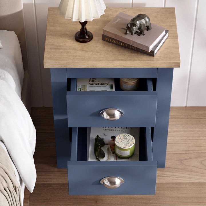 [Blue] Bedside table top view with both drawers open, lamp, books, decorative jars, bed edge, and wood floor