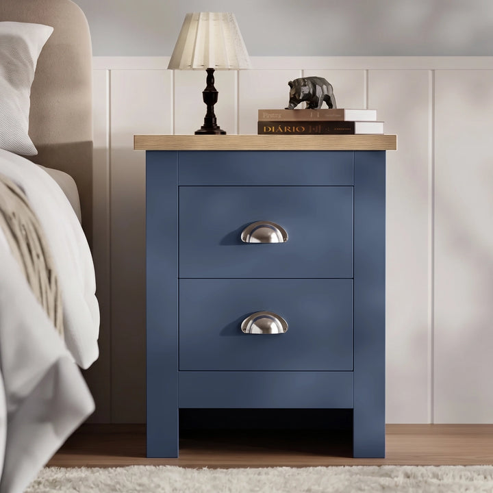 [Blue] Bedside table front view beside a bed, with lamp, stacked books, oak top, panelled wall, and rug