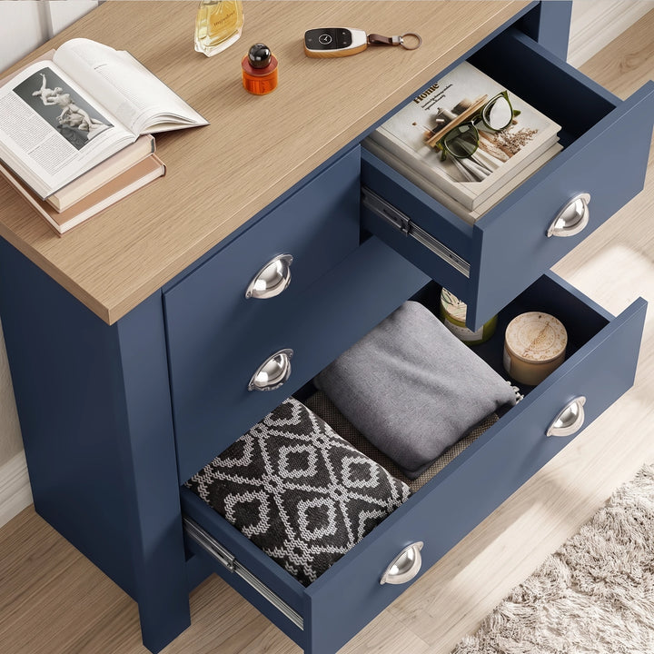 [Blue] Chest of drawers top angle with two drawers open, folded textiles, books, small bottles, and oak surface
