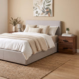 [Dark Bronx] modern bed with a light gray upholstered frame and headboard, dressed in white and beige bedding, styled in a warm neutral bedroom with a dark wood nightstand, woven rug, and botanical wall art.