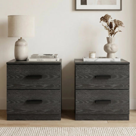 [Black] Pair of two drawer bedside tables placed side by side with ceramic lamp and stacked books on left and sculptural vase with dried flowers on right