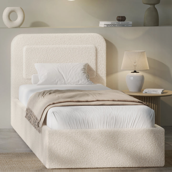 Beige boucle single ottoman bed angled view, rounded pill headboard beside fluted lamp and pebble vase shelf