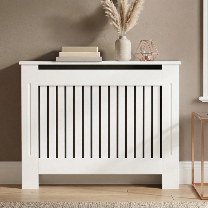 [Medium] Chelsea White Radiator Cover angled three-quarter view in taupe room on wooden floor with jute rug and rose gold accessories on top shelf