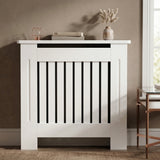 [Small] Chelsea White Radiator Cover front-facing in warm taupe room with rose gold lantern, stacked books, and pampas grass vase on top shelf