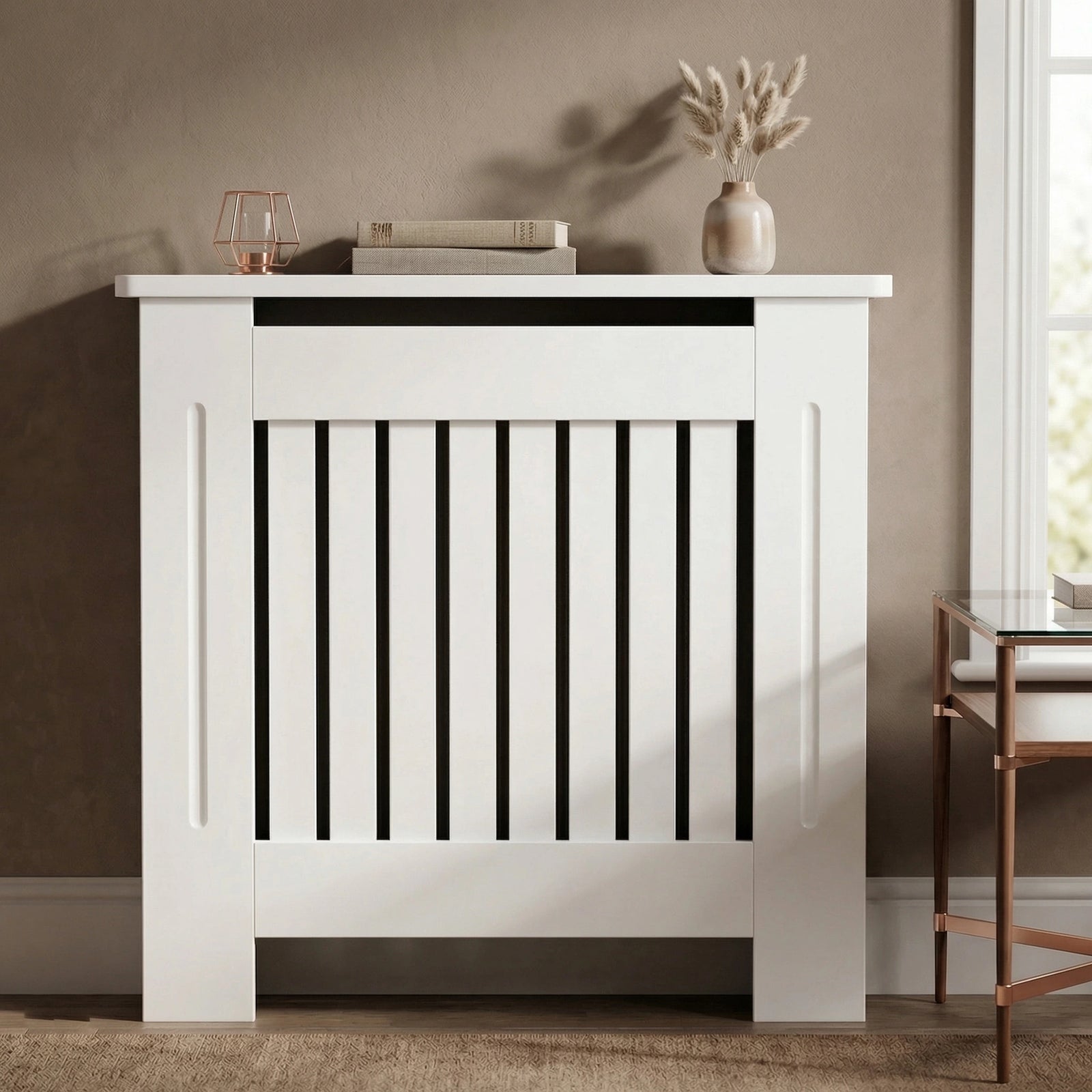 [Small] Chelsea White Radiator Cover front-facing in warm taupe room with rose gold lantern, stacked books, and pampas grass vase on top shelf