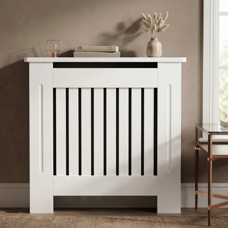 [Small] Chelsea White Radiator Cover front-facing in warm taupe room with rose gold lantern, stacked books, and pampas grass vase on top shelf