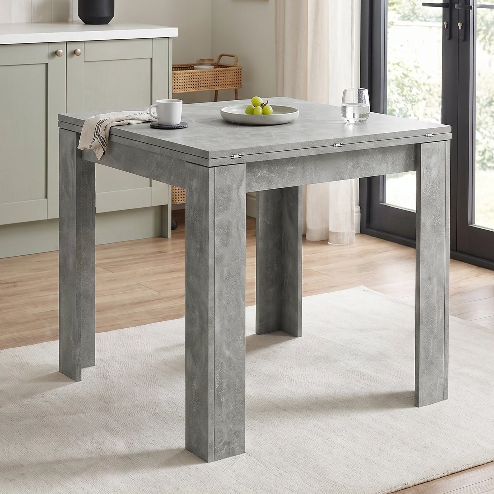 [Concrete] Closed square flip-top dining table styled with a mug, fruit bowl and glass in a bright kitchen.