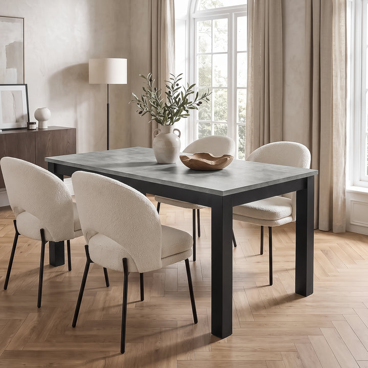 [140cm | Concrete] Closed 140cm dining table with four cream chairs, concrete top, leafy vase, wooden bowl and black frame.
