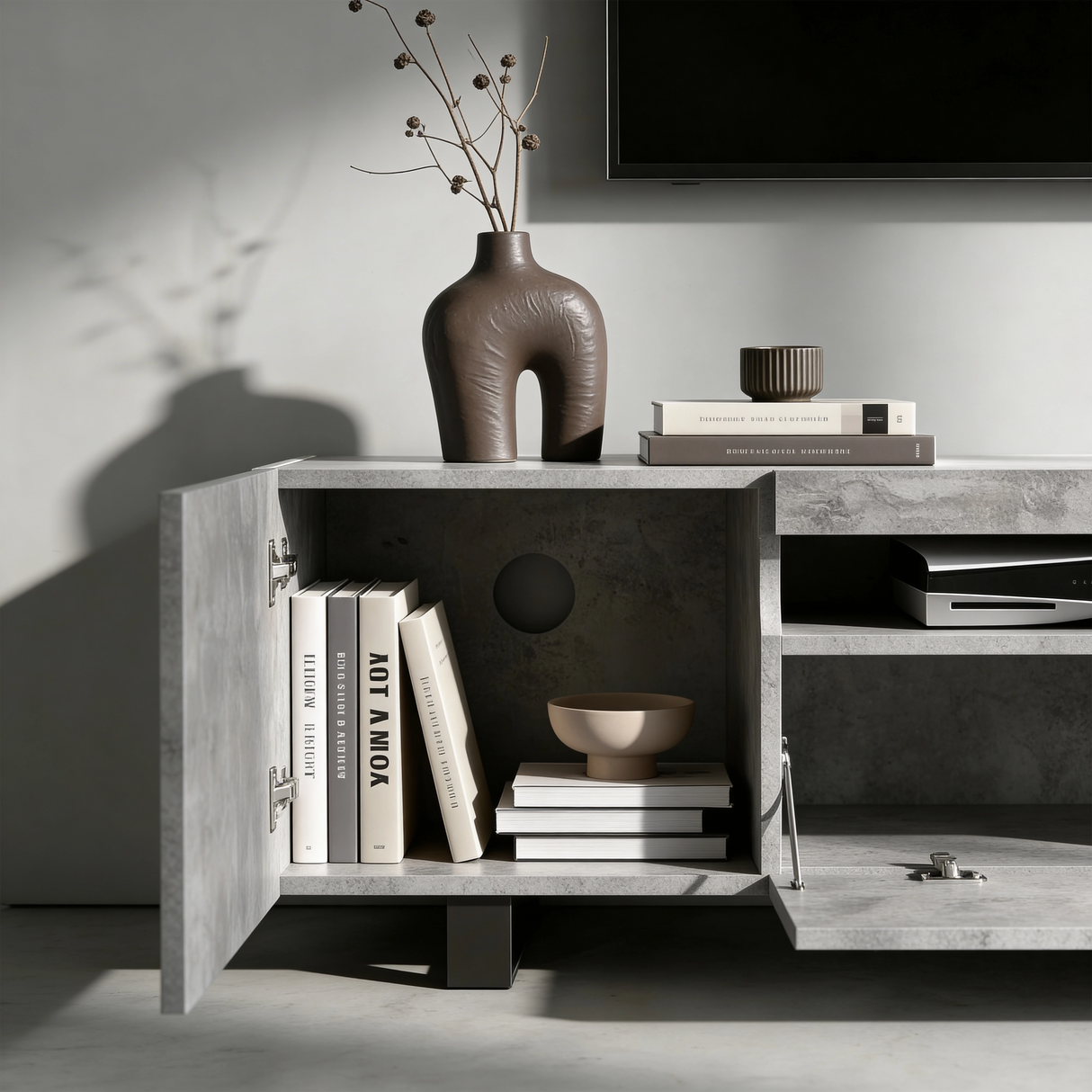 [Concrete] Kystra TV stand left cabinet door open close-up showing internal shelf with books and a ceramic bowl, chrome hinge hardware, round cable routing hole, and AV equipment visible in the centre shelf
