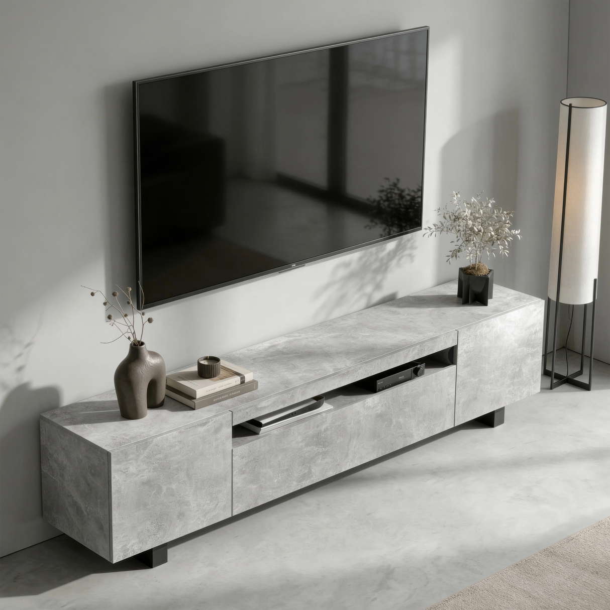 [Concrete] Kystra TV stand viewed from above at an angle in a minimal living room, showing the concrete-effect top surface, open centre shelf with AV unit, dark vase on the left, and floor lamp to the right
