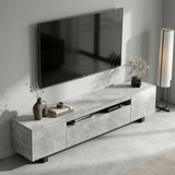 [Concrete] Kystra TV stand viewed from above at an angle in a minimal living room, showing the concrete-effect top surface, open centre shelf with AV unit, dark vase on the left, and floor lamp to the right
