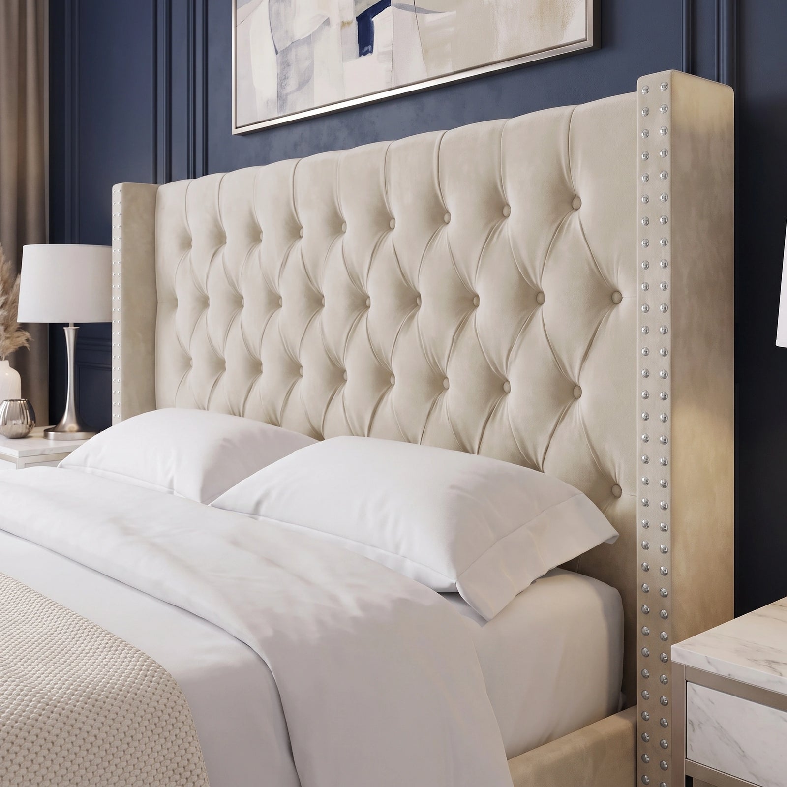 [King | Beige]A large upholstered headboard in a warm cream or beige fabric, featuring deep button tufting and nailhead trim on the side rails, shown on a bed with white bedding in a styled bedroom.