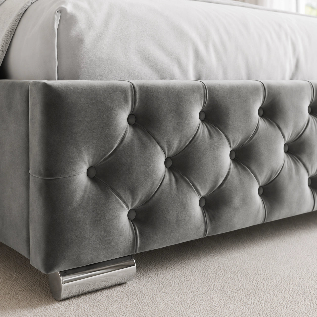 [Double | Grey] A close-up of a gray tufted velvet bed frame with a metallic silver footrest, set against a light gray carpet and white bedding.