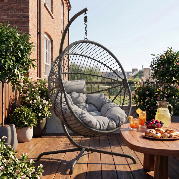 [Dark Grey] Hanging egg chair on a wooden deck with a table of snacks and drinks, surrounded by plants and a building in the background.