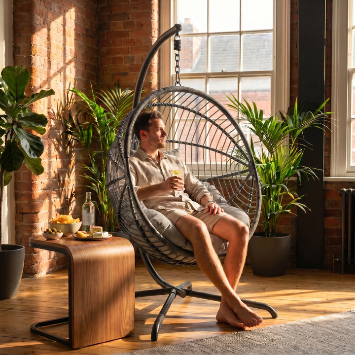 [Dark Grey] Man relaxing in a hanging egg chair in a sunlit room with plants and a wooden table