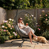 [Dark Grey] Woman sitting in a hanging chair in a garden with flowers and a dog