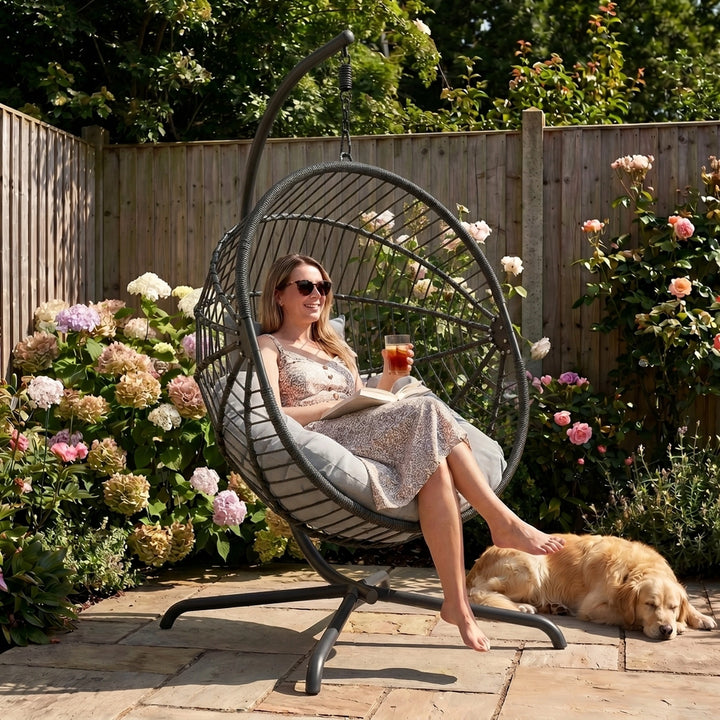 [Dark Grey] Woman sitting in a hanging chair in a garden with flowers and a dog.
