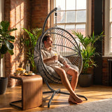 [Dark Grey] Man relaxing in a hanging egg chair in a sunlit room with plants and a wooden table
