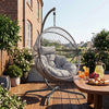 [Grey] Hanging chair on a wooden deck with a table of snacks and drinks, surrounded by plants and a building in the background