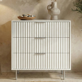 White Flutrina 3 drawer chest with vertical panels and slim metal legs in a bedroom setting