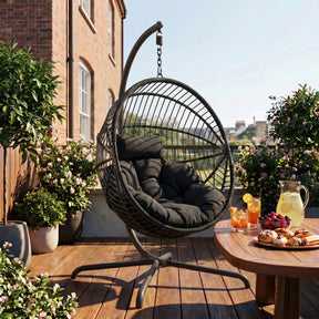 [Black] Hanging chair on a wooden deck with a table of snacks and drinks, surrounded by plants and a building in the background