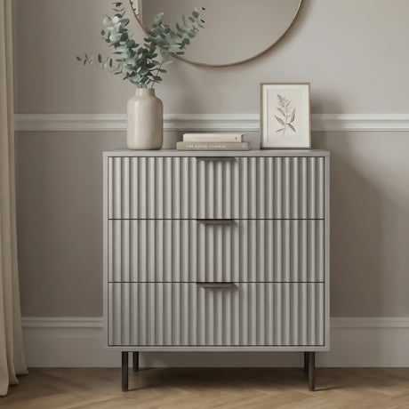 Grey chest of drawers with three storage drawers and flutrina front detailing placed in a bedroom