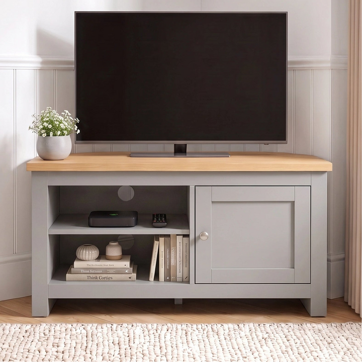 [Grey] Corner TV stand with a natural wood top, shown in a styled room setting with a black flat-screen TV on top, a small potted plant, and decorative items on the open shelf.