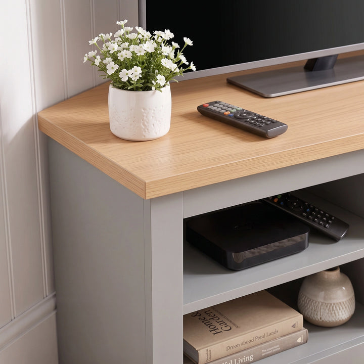 [Grey] Corner TV unit with a grey base, styled with a potted white flower, a TV remote, and a television on top, set against a white paneled wall.