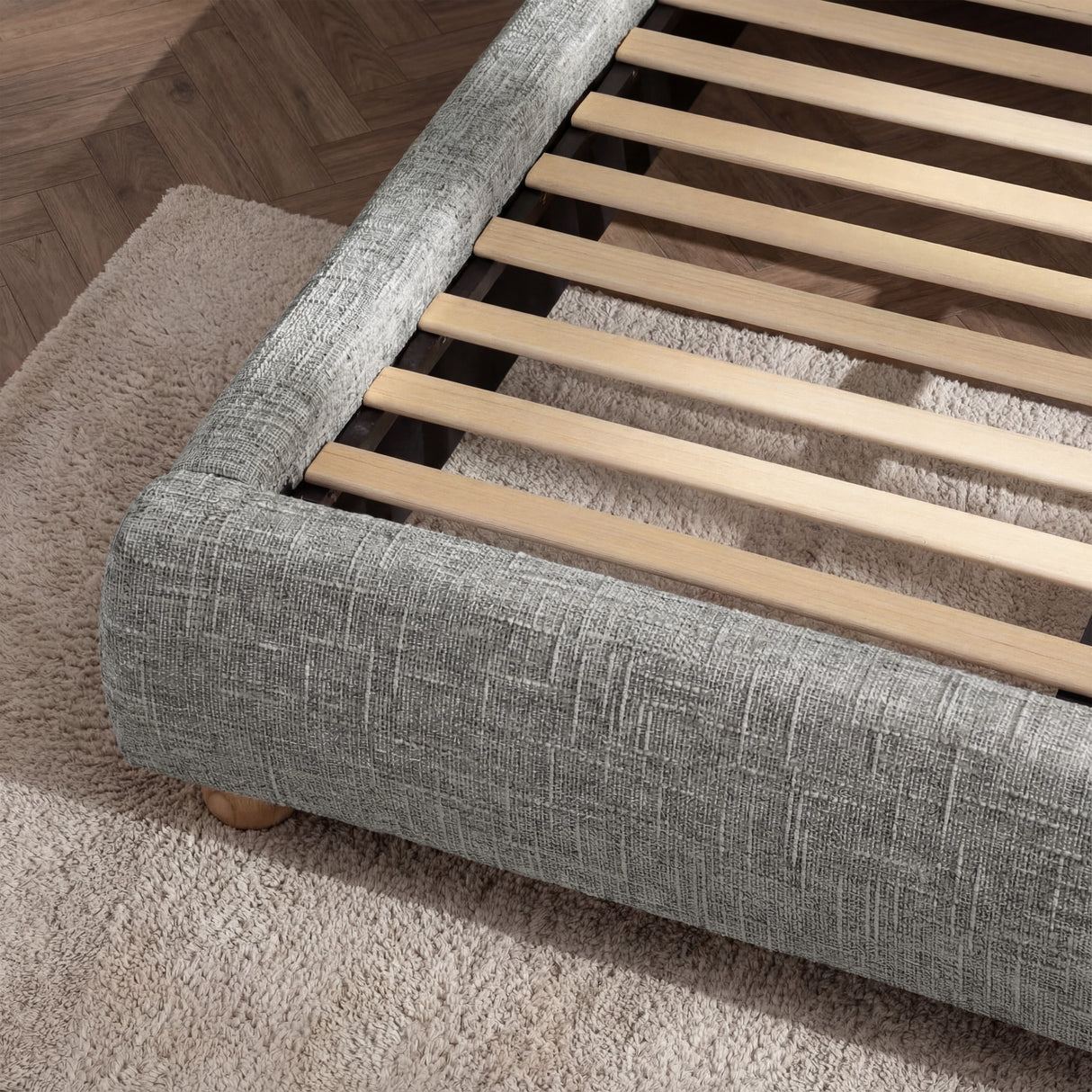 Angled view of a grey fabric-wrapped upholstered bed base corner with exposed wooden slats