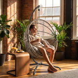[Grey] Man relaxing in a hanging egg chair in a sunlit room with plants and a wooden table